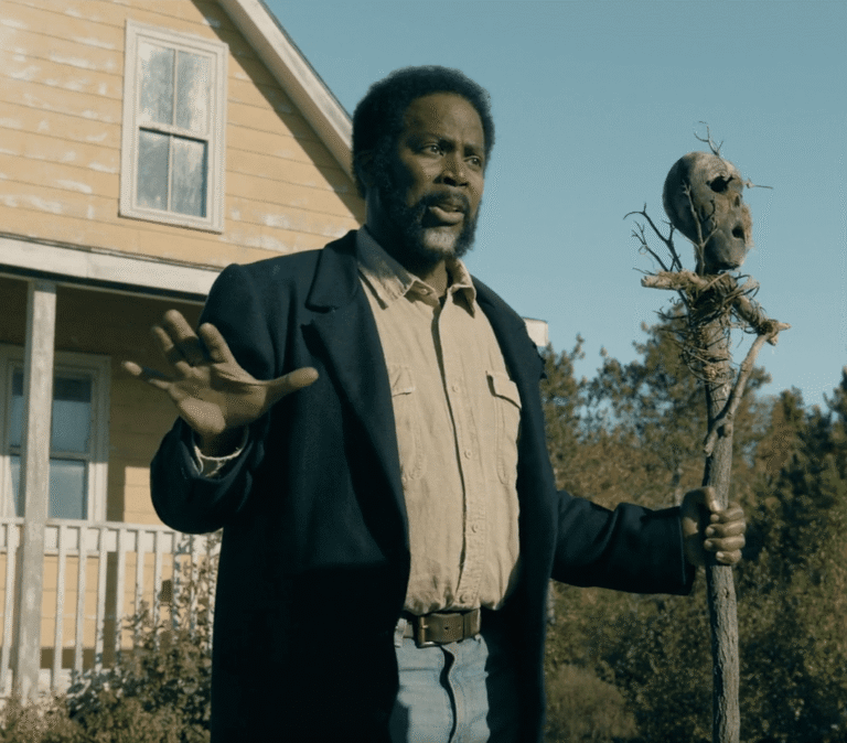 A FROM Season 4 Episode 7 scene showing a character holding a totem outside a yellow house while discussing Boyd’s plan.