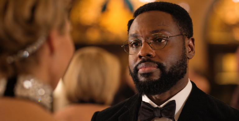 Leighton wearing round glasses and a black tuxedo, facing a woman in a warmly lit room in Dark Matter Season 2 Episode 6.