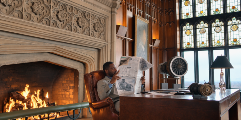 Leighton reading a newspaper beside a fireplace, with turbine-shaped objects and stained-glass windows around his desk.